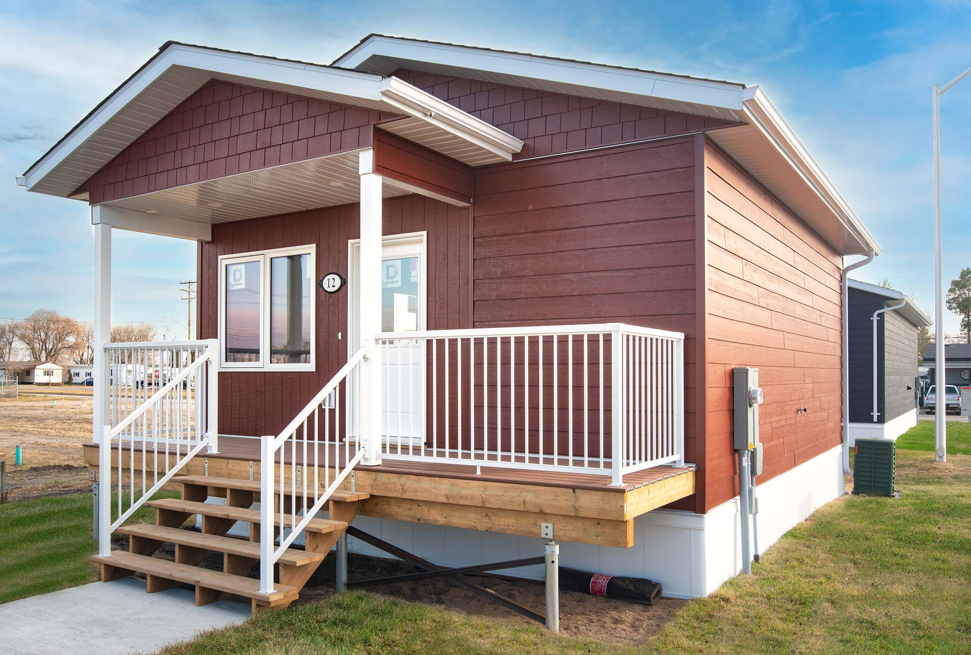 Exterior view of the front of the Bluebell small home style at Glendale Homes Park in Brandon, Manitoba. The home has front porch with white metal railing. The home has red siding and white trim.