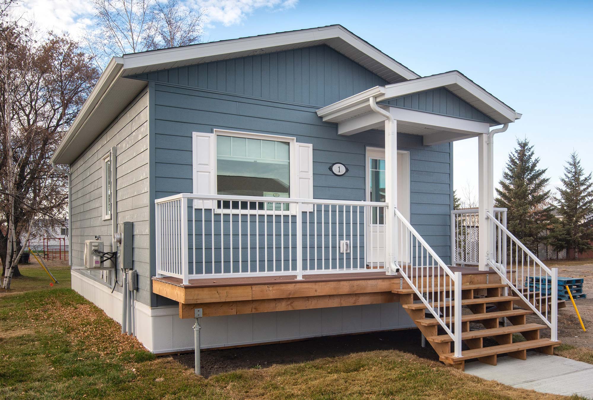 Exterior view of the front of the Daisy small home style at Glendale Homes Park in Brandon, Manitoba. The home has front porch with white metal railing. The home has medium blue siding and white trim.