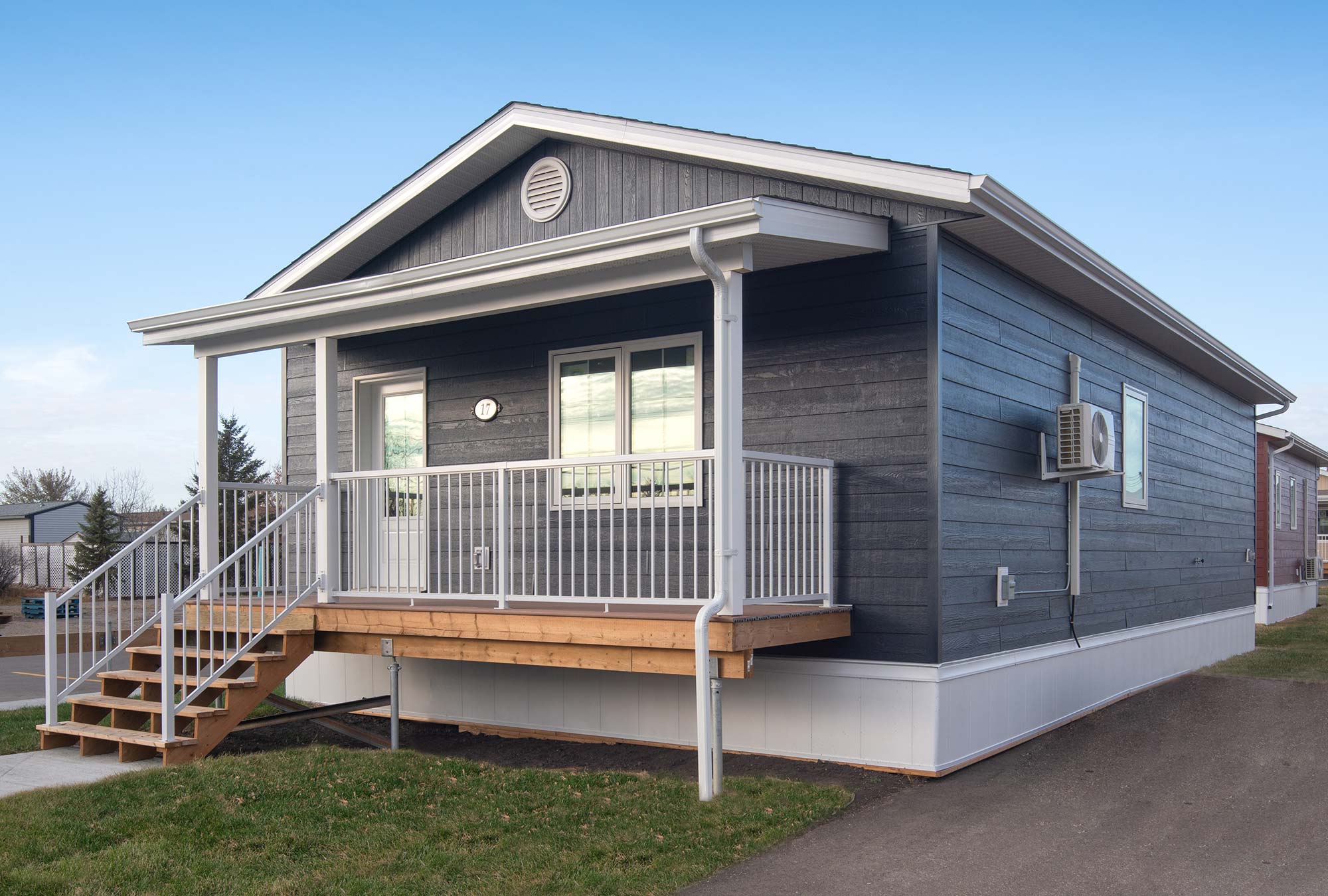 Exterior view of the front of the Wild Rose small home style at Glendale Homes Park in Brandon, Manitoba. The home has front porch with white metal railing. The home has medium blue siding and white trim.